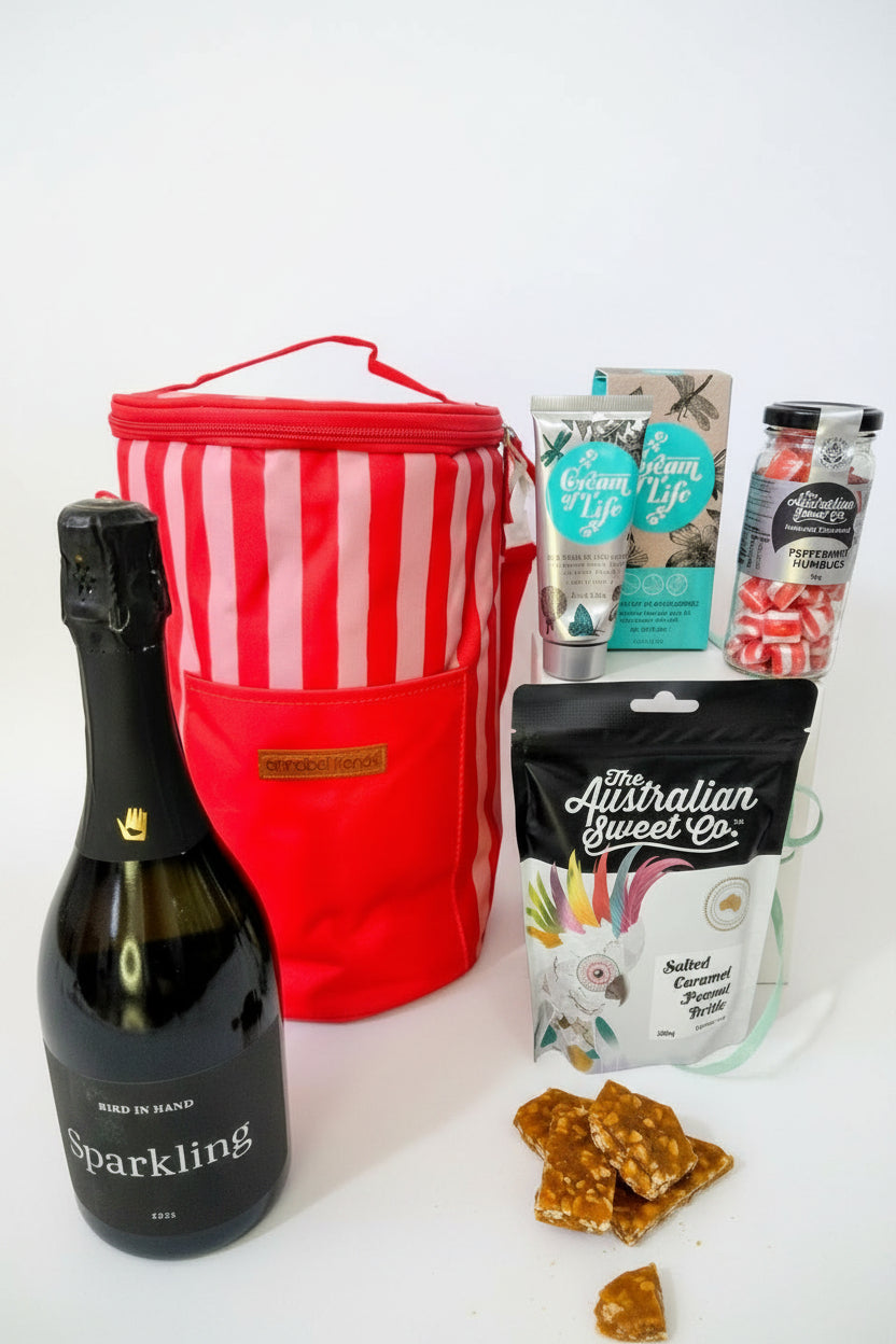 Bottle of sparkling wine, red striped cooler bag, and assorted snacks on a textured surface. Gifts for her