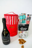 Bottle of sparkling wine, red striped cooler bag, and assorted snacks on a textured surface. Gifts for her